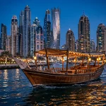 Traditional wooden dhow boat lit with warm lights sailing on Dubai Marina at dusk with the illuminated skyline behind