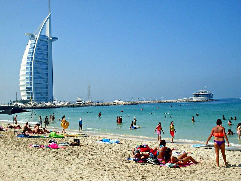 Beachgoers swimming and sunbathing at a Dubai public beach with the Burj Al Arab behind