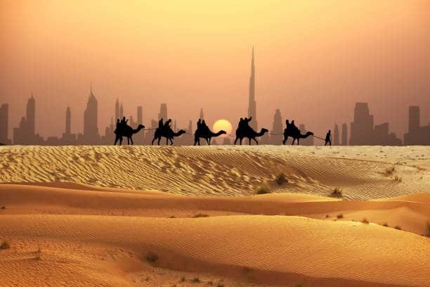 A camel caravan crossing desert dunes at sunset with the Dubai skyline far behind